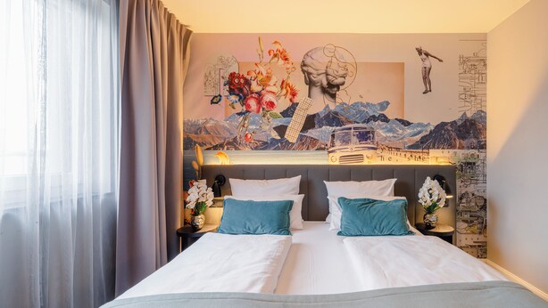 A hotel room with a bed, a nightstand, a lamp, and a wall mural. - PLAZA INN Ulm (Ulm)