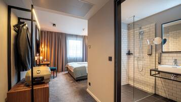 Ein Hotelzimmer mit Bett, Schreibtisch mit Lampe, Koffer, Duschbereich mit Glastür sowie Bad mit Waschbecken und Spiegel.