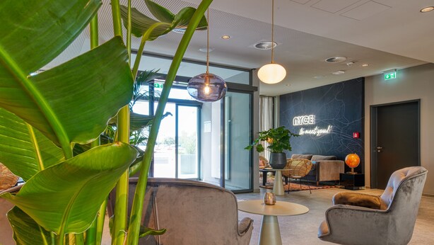 A modern lobby with a round table, two grey armchairs, and large green plants. - PLAZA INN Ulm (Ulm)