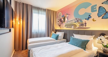 Habitación de hotel con dos camas, un ventanal con cortinas, un mural en la pared con un autobús y mariposas, y una mesita de noche con un jarrón de flores.