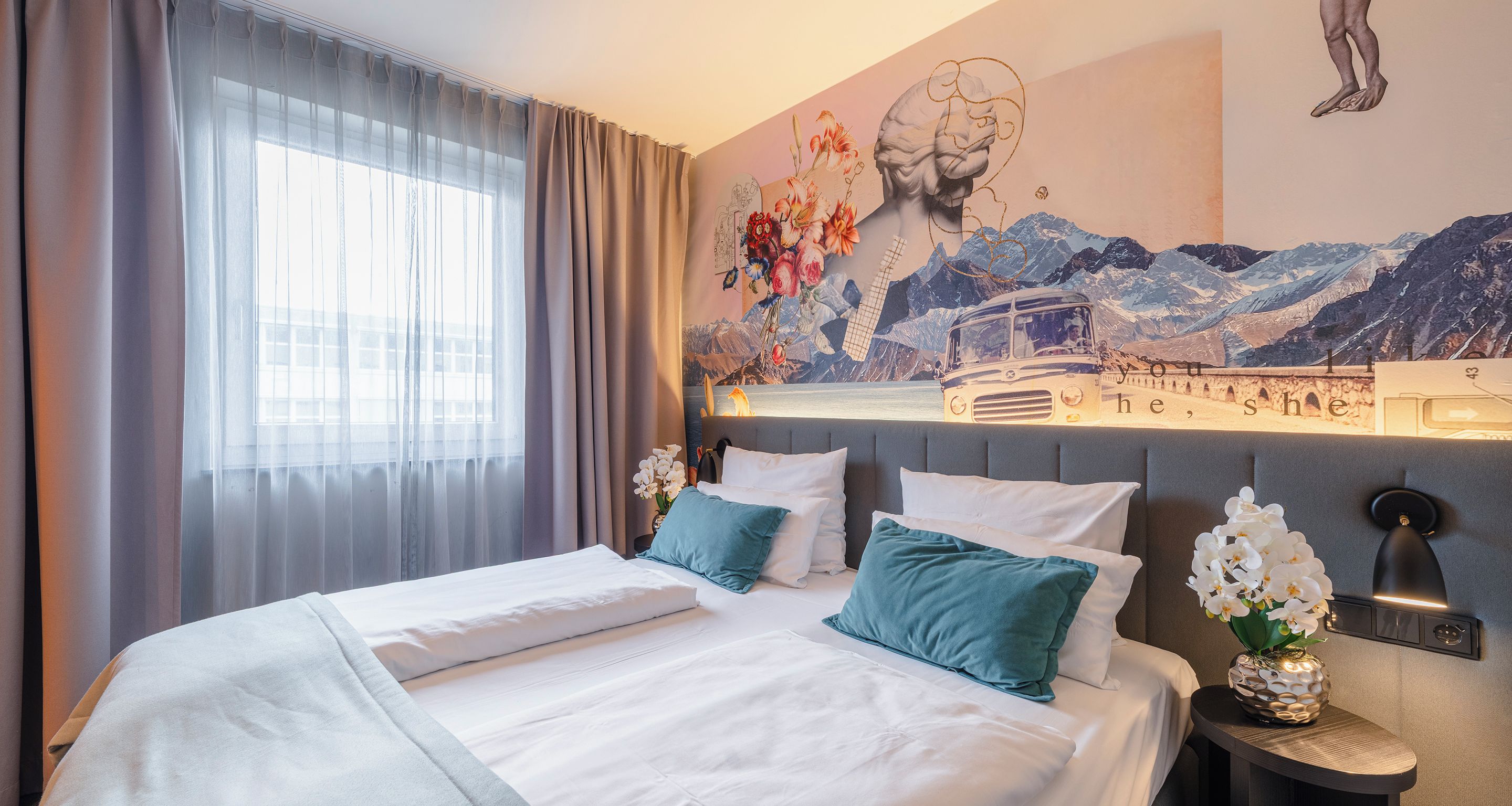 Una habitación de hotel con cama, mesita de noche, lámpara de pared y ventana con cortinas.