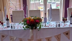 A table set for a wedding reception with a floral centerpiece, table number sign, and a banner with initials. - The Castle Inn (North Berwick)