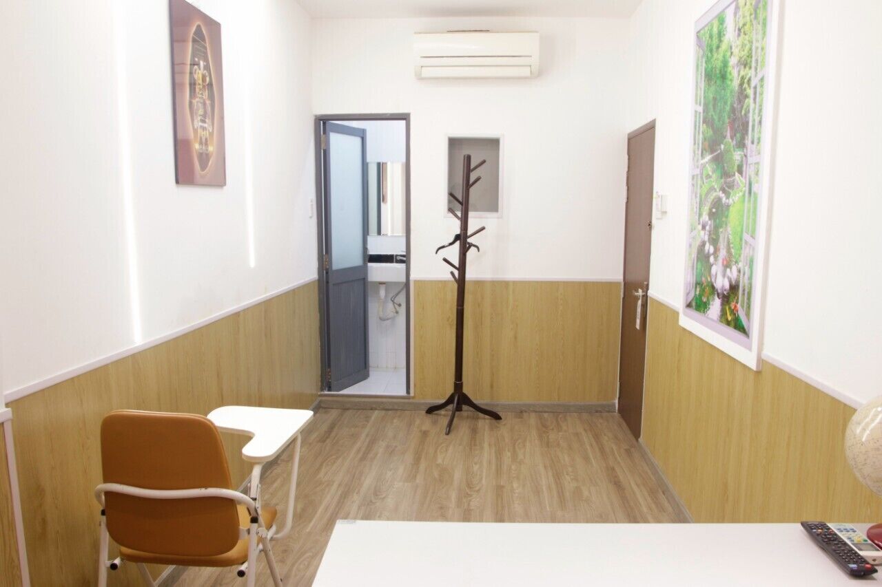 A small, well-lit room with a wooden floor, a white desk, a brown chair, a coat rack, and a door leading to another room.