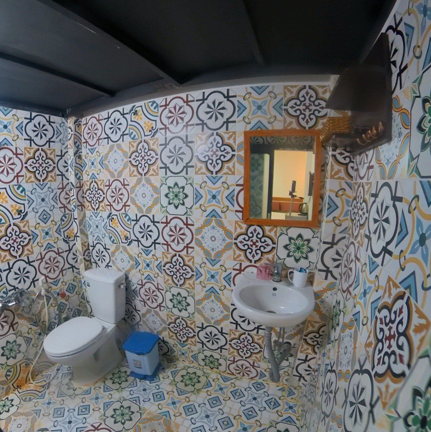 Une salle de bain avec des toilettes, un lavabo et un miroir. Les murs sont recouverts de carreaux colorés à motifs.