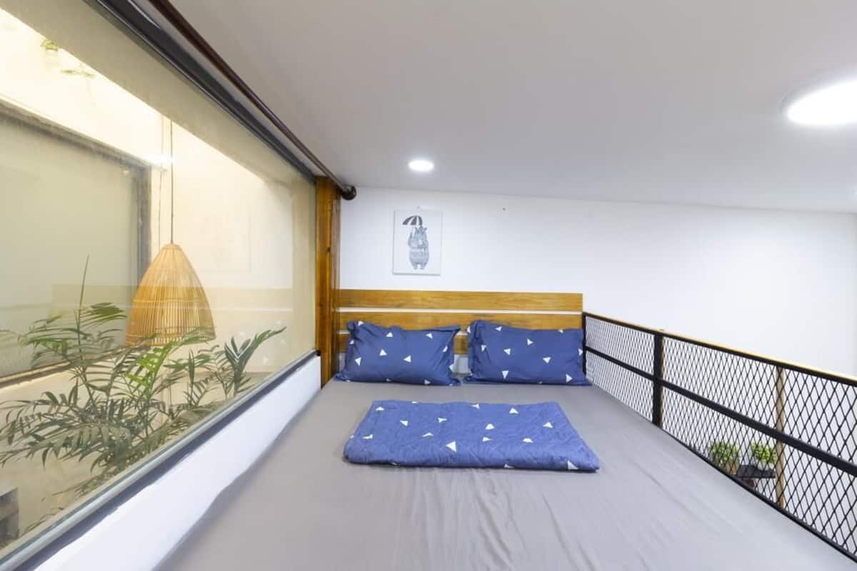 A single bed with a wooden headboard, a blue blanket, and matching pillows. There is a window on the left side with a view of greenery outside.