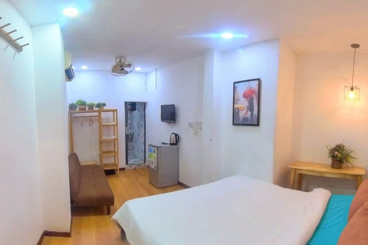 A small, well-lit room with a bed, a desk, a chair, a small cabinet, a TV, and a plant.