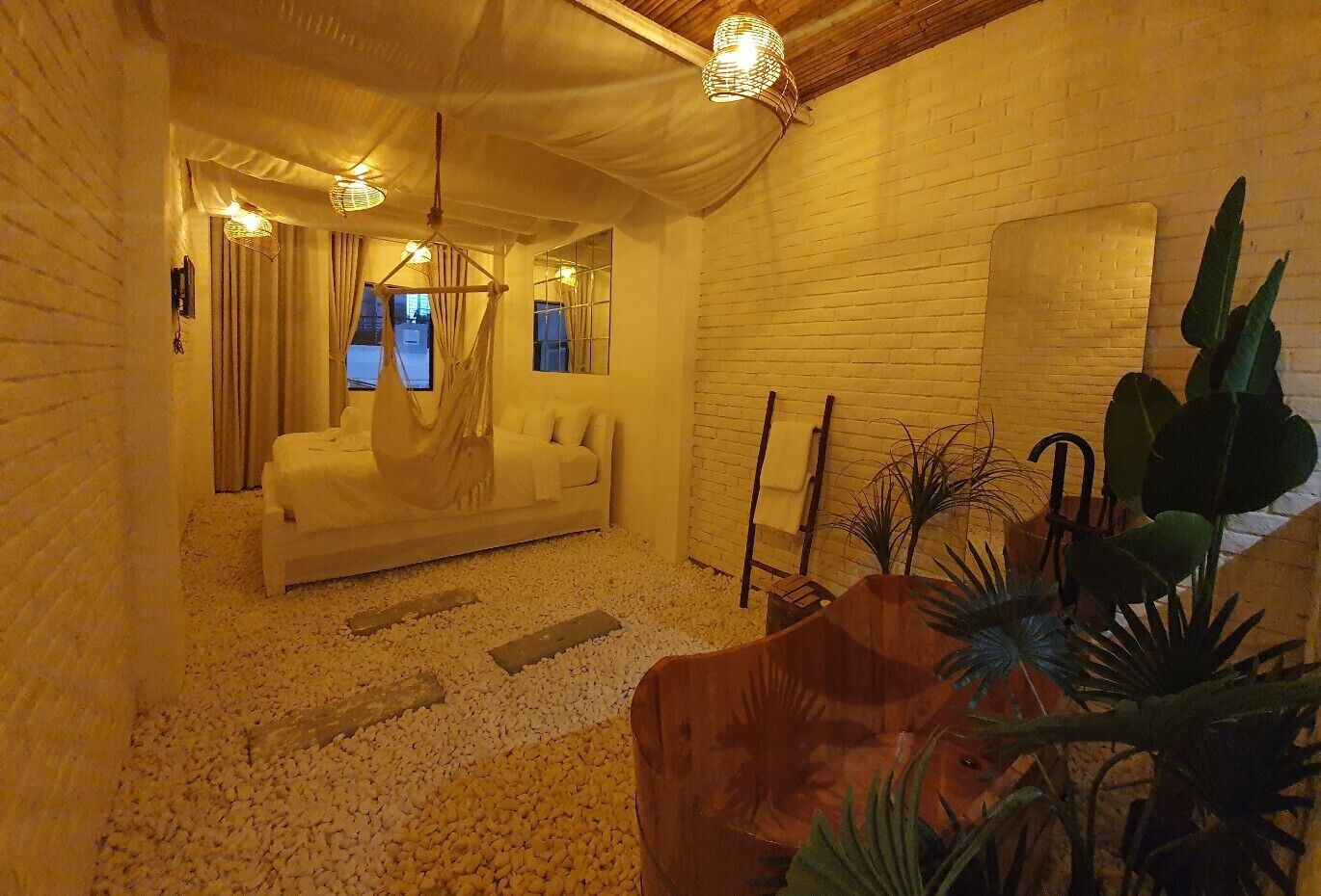 A cozy room with a white sofa, a hanging chair, and a small wooden table. There are plants and a brick wall in the background.