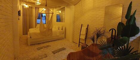 A cozy room with a white sofa, a hanging chair, and a small wooden table. There are plants and a brick wall in the background.