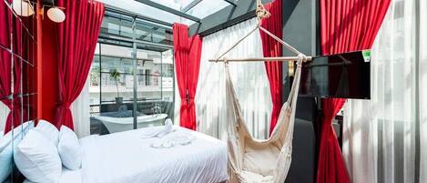 A modern hotel room with a large bed, red curtains, a hammock, and a TV.