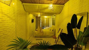 A narrow hallway with a white brick wall, a wooden ceiling, and a sofa with a yellow blanket. There are large green plants and a mirror on the right side. - Language Exchange Hotel 3 (Ho Chi Minh City)