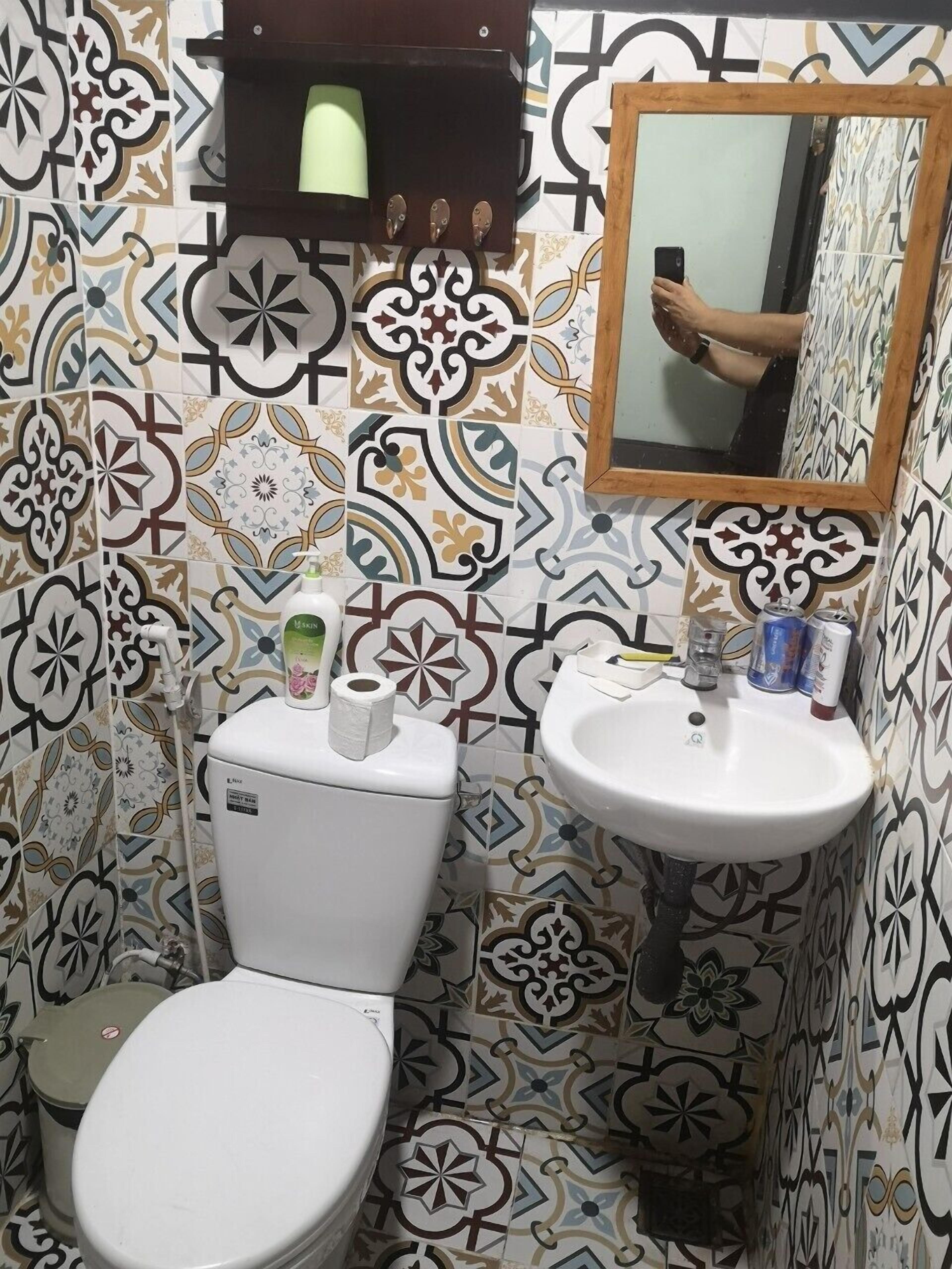 A bathroom with a toilet, sink, and mirror. There is a patterned tile backsplash and a small shelf above the toilet.