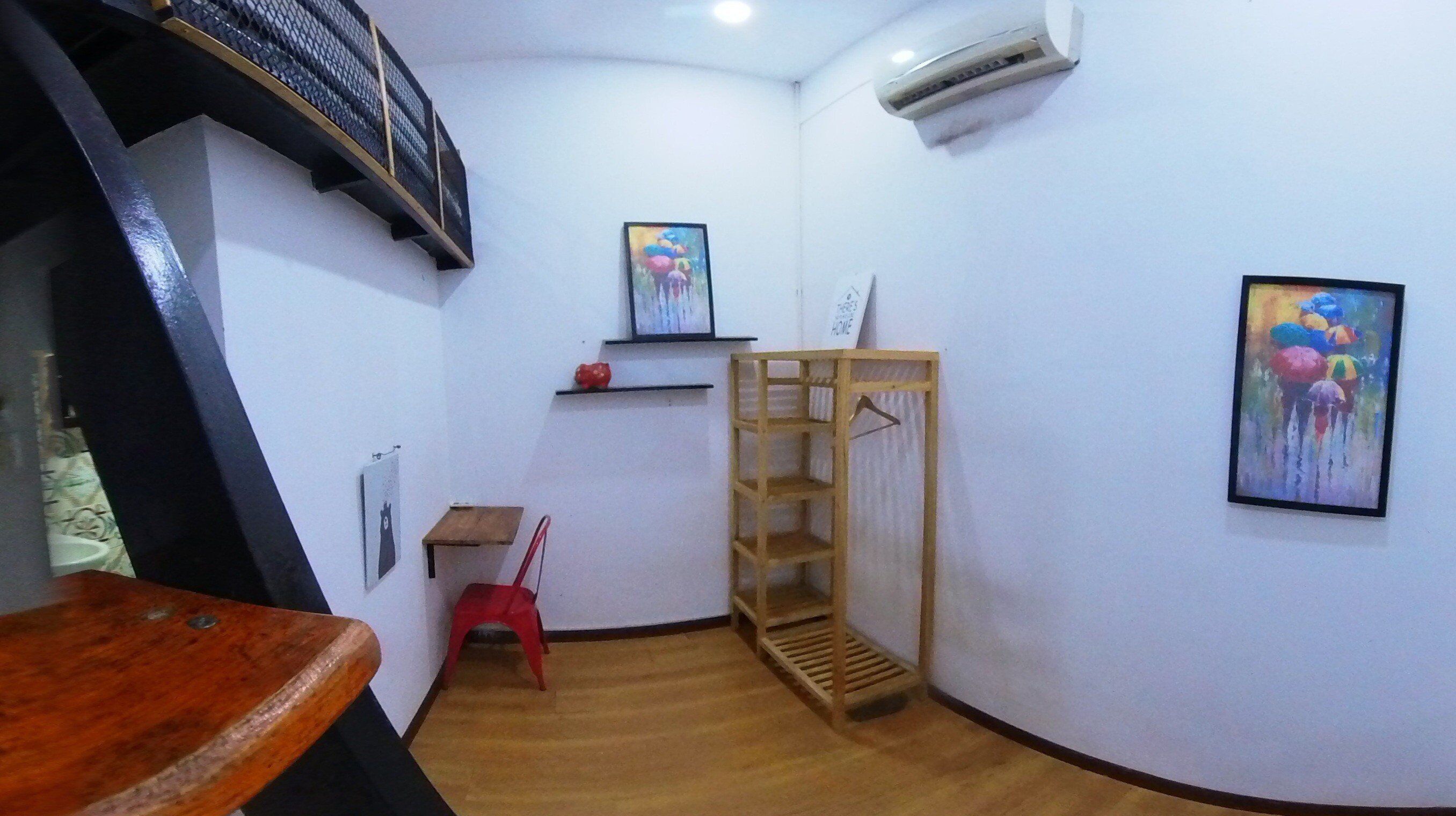 Una habitación pequeña y estrecha con una mesa de madera, una silla de plástico rojo, un estante de madera y dos cuadros en las paredes.