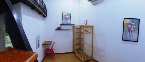 Una habitación pequeña y estrecha con una mesa de madera, una silla de plástico rojo, un estante de madera y dos cuadros en las paredes.