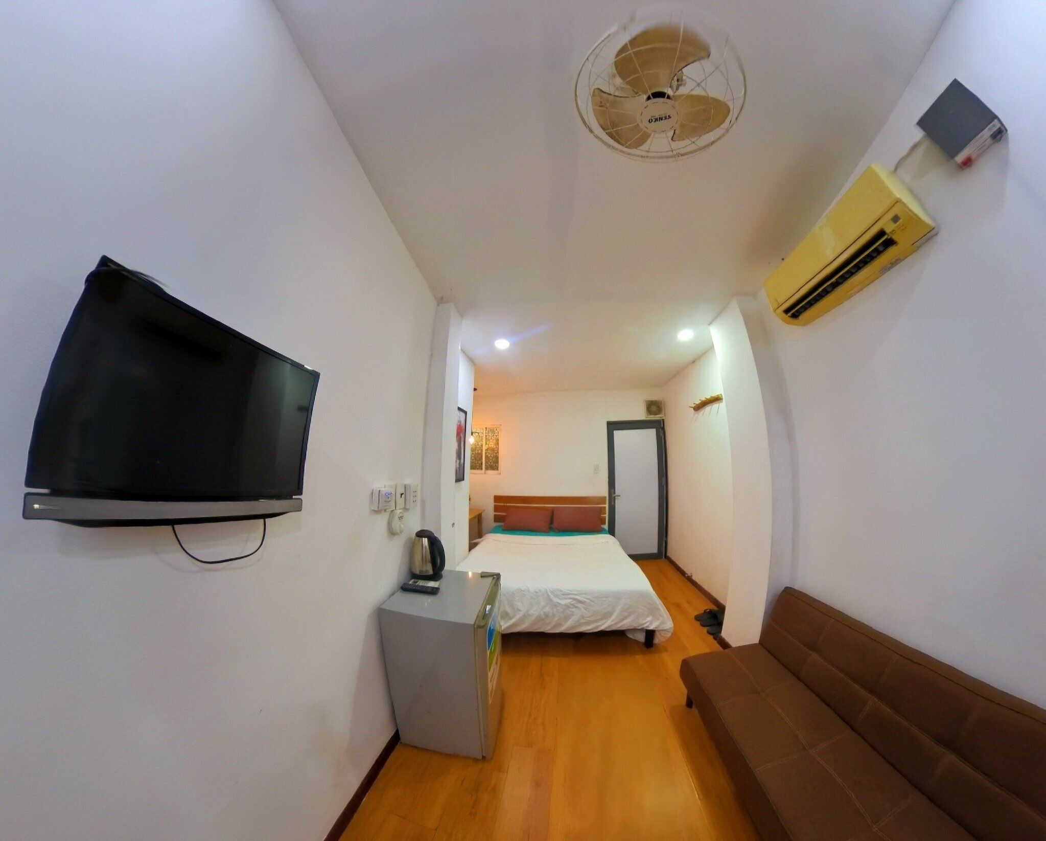 A narrow hotel room with a bed, a small desk, a television, a ceiling fan, and an air conditioning unit.