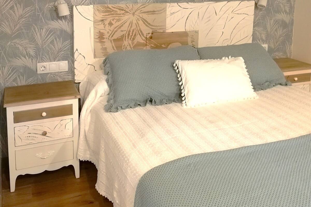 A bedroom with a wooden ceiling, a bed with a blue and white blanket, a nightstand with a lamp, and a wall with a leaf pattern.