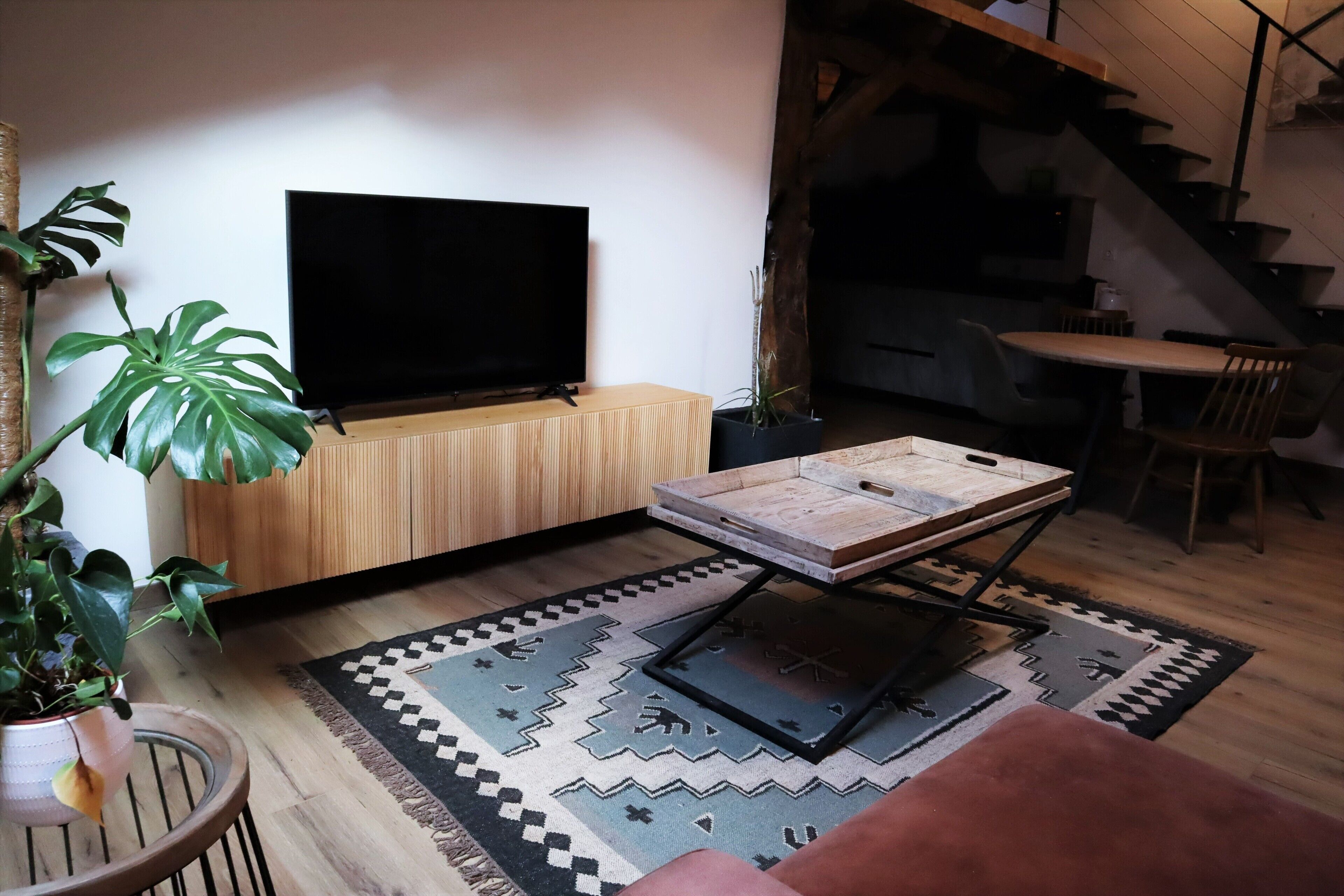 A modern living room with a flat-screen TV, a wooden cabinet, a dining table, and a staircase leading to an upper level.