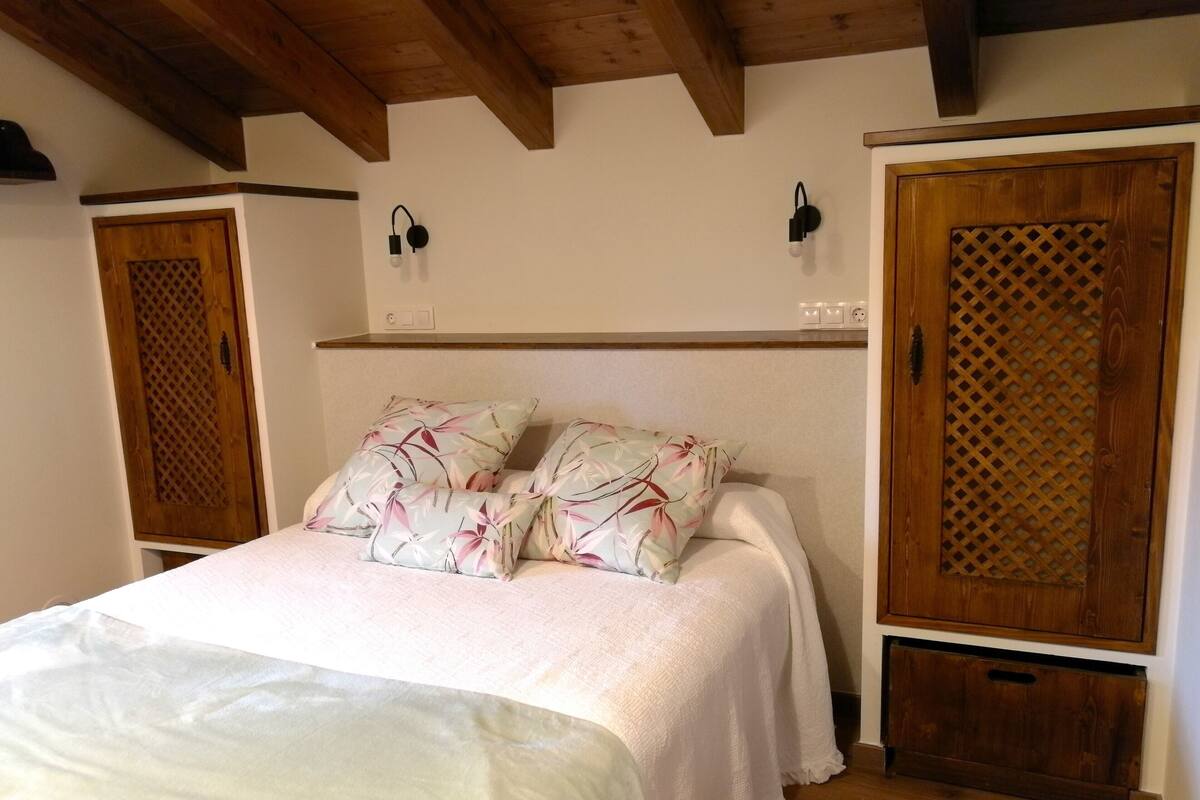 A bedroom with a wooden ceiling, a bed with floral pillows, a white bedspread, two wall-mounted light fixtures, and a wooden wardrobe with a lattice design.