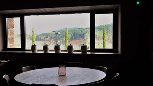A round table with six chairs and a vase in the center, set against a window with a view of trees and hills. - Eguen Goiko (Ispaster)