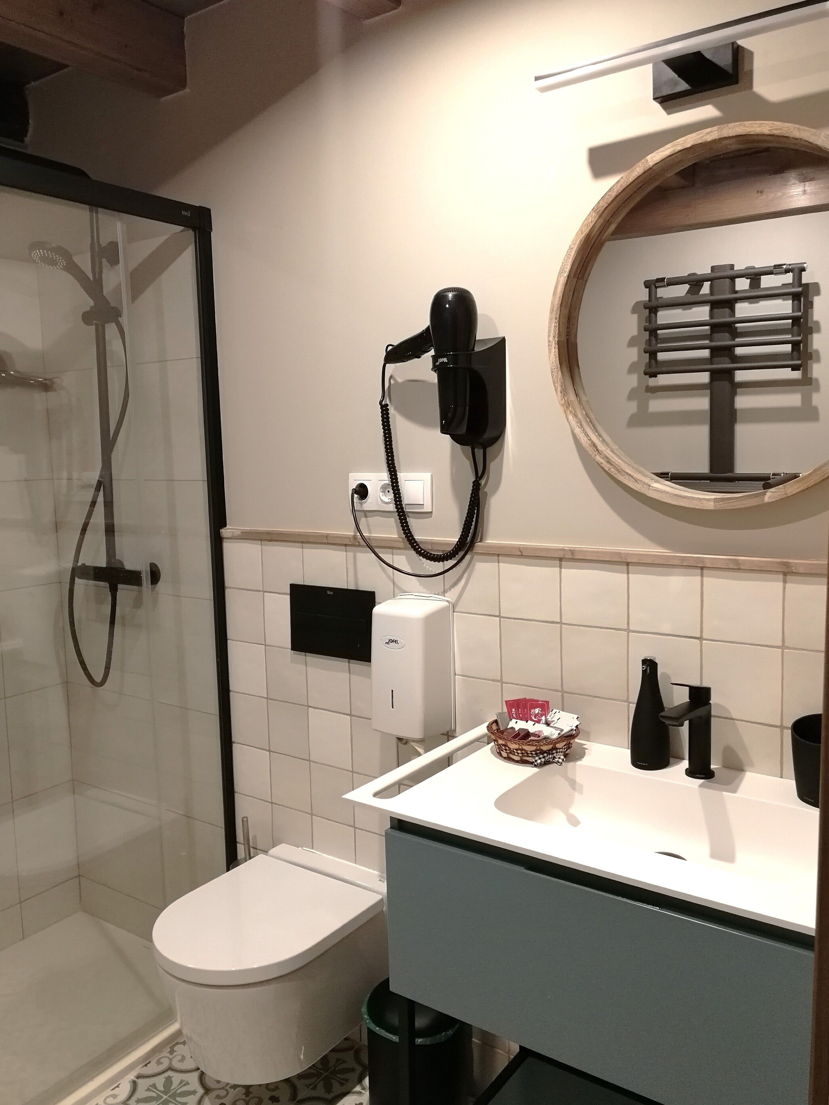 A modern bathroom with a glass shower enclosure, a toilet, a sink with a towel rack, and a soap dispenser.