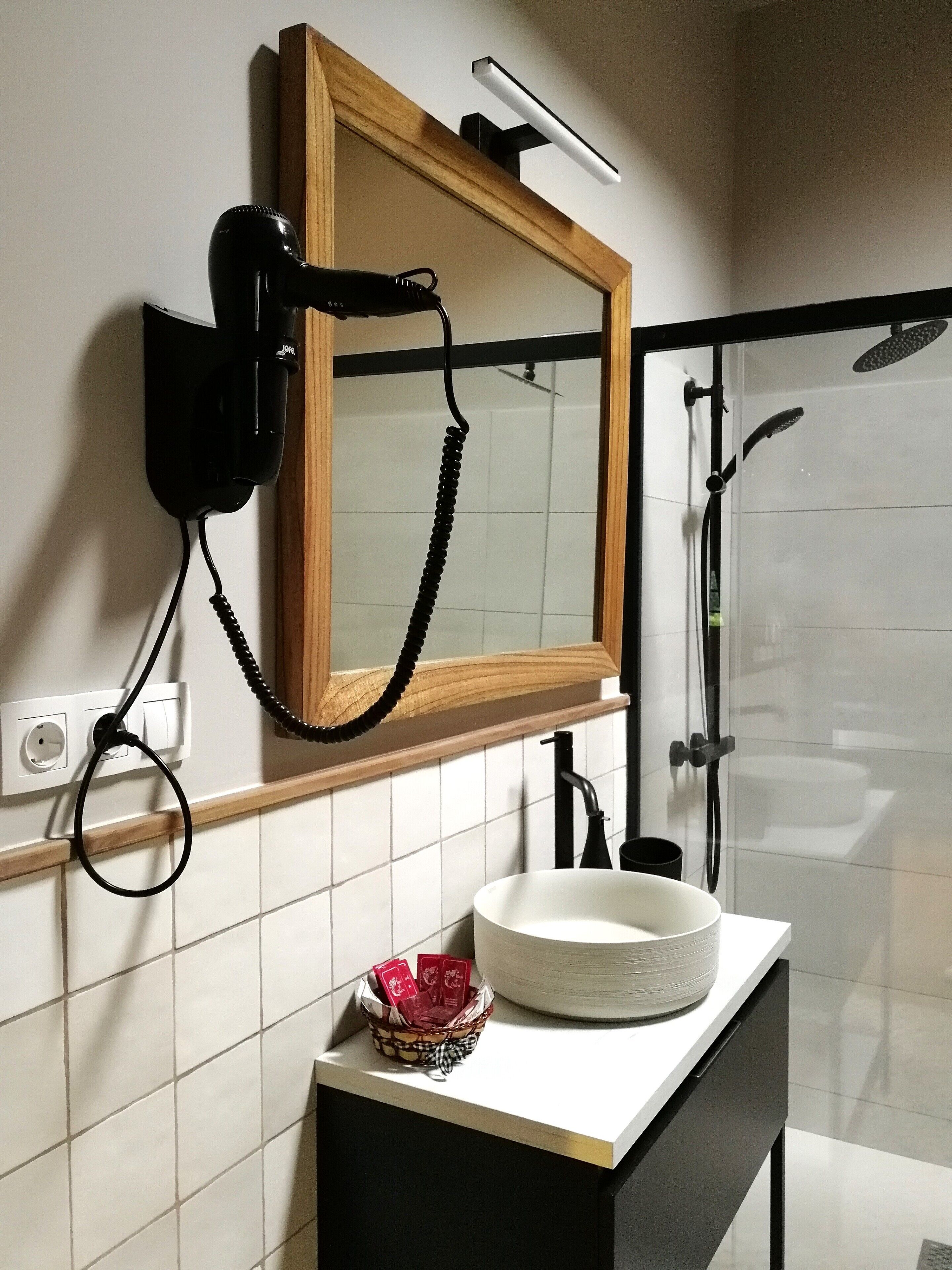 Une salle de bain avec un miroir encadré en bois, un sèche-cheveux noir, un lavabo blanc avec un bac blanc, et un espace douche doté d’une porte en verre.
