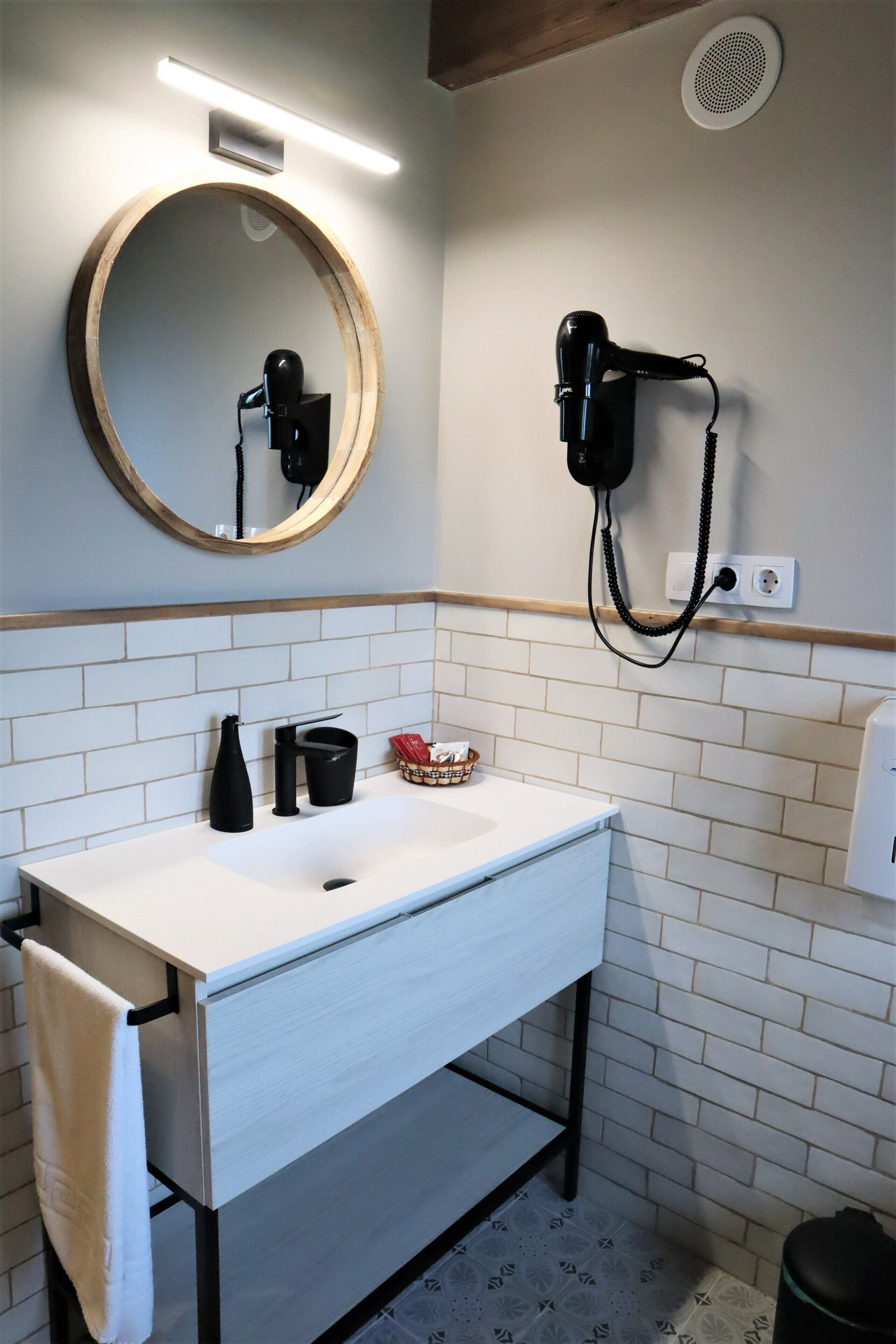 Een badkamer met een wit wastafel, een ronde spiegel, een haardroger en een handdoek aan de muur.