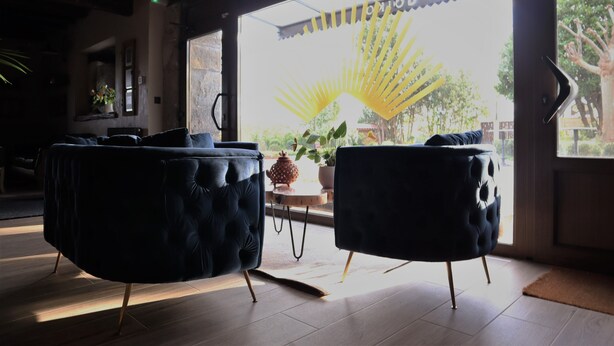 A room with two dark upholstered chairs, a wooden floor, and a large window with a view of a garden and a sign reading "GUEVEN OYUN". - Eguen Goiko (Ispaster)