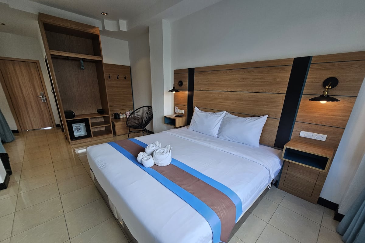 A hotel room with a large bed, wooden headboard, bedside tables, a wardrobe, and a desk with a chair.