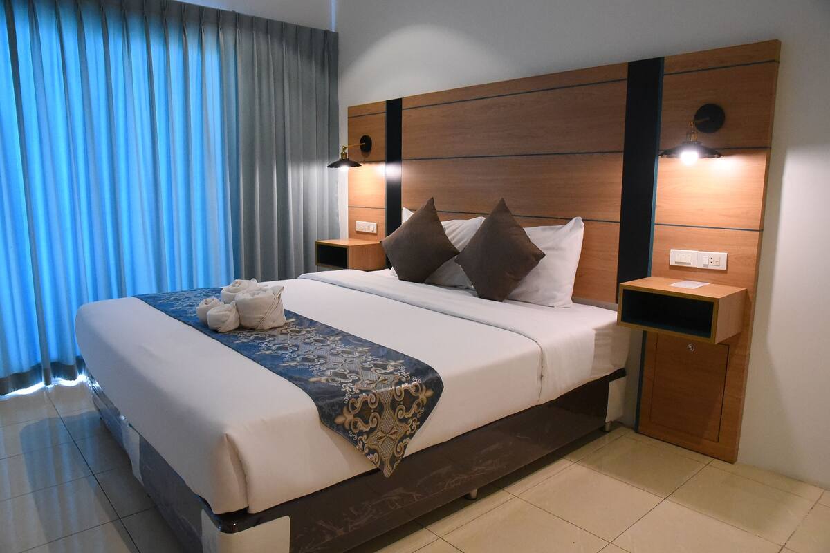 A neatly made bed with a blue and white bedspread, two brown pillows, and a decorative throw. There are two bedside tables with lamps, and a window with curtains.