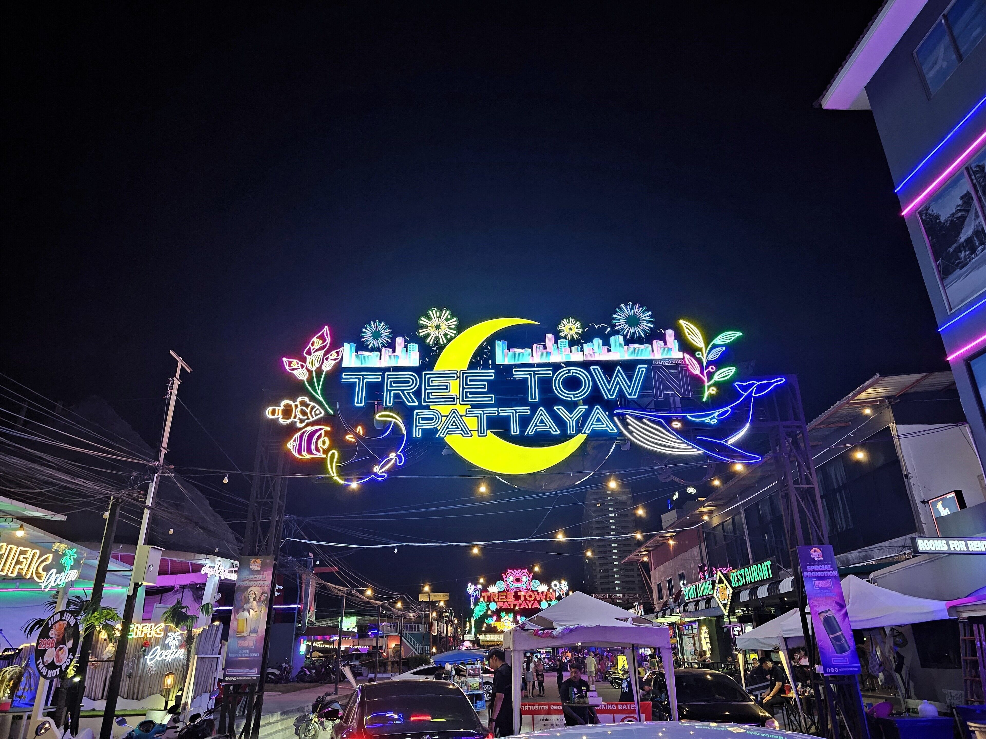 A brightly lit street with a neon sign reading "TREE TOWN PATAYA" and various shops and restaurants.