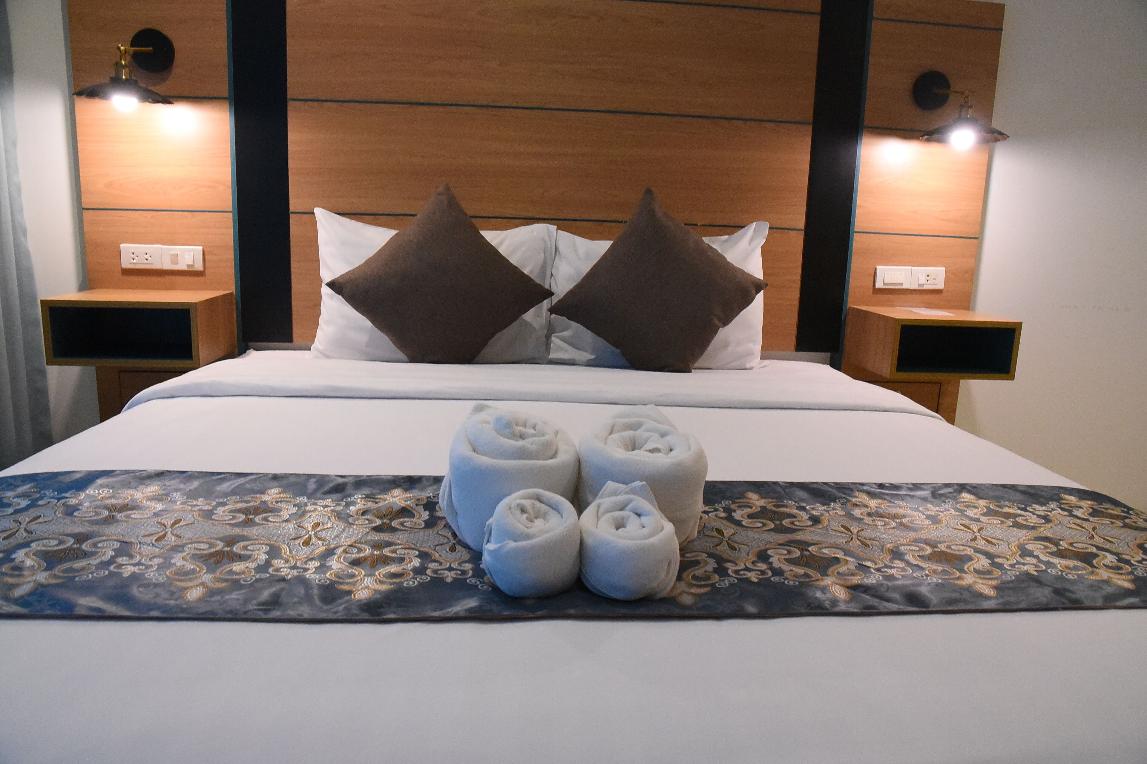 A neatly made bed with white linens, two brown pillows, and four rolled towels on a patterned bedspread.