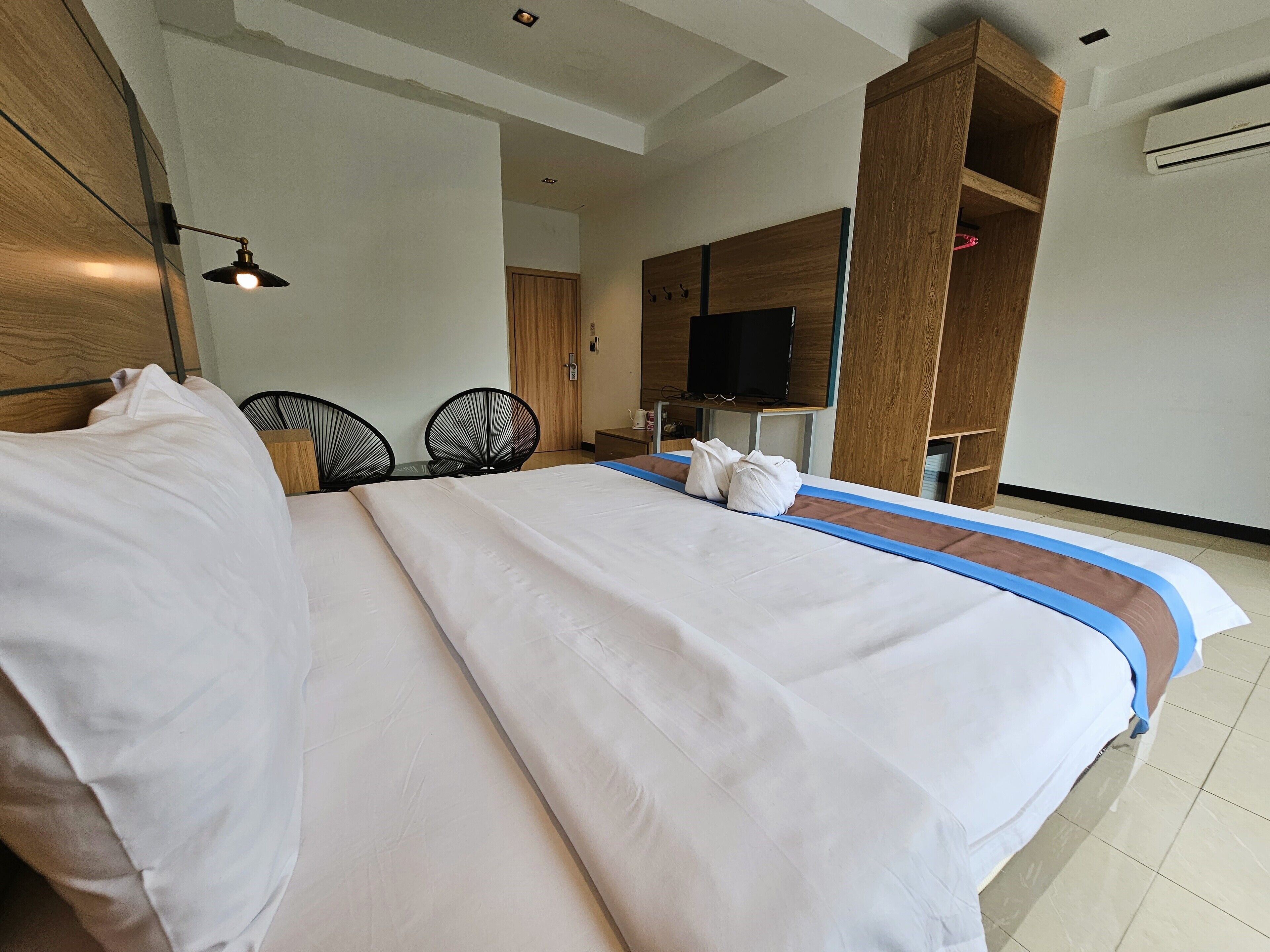 A neatly made bed with white linens, a wooden headboard, and a blue accent on the bed's edge. There is a television mounted on a wooden panel above the bed, and a wooden wardrobe with a mirror to the right.