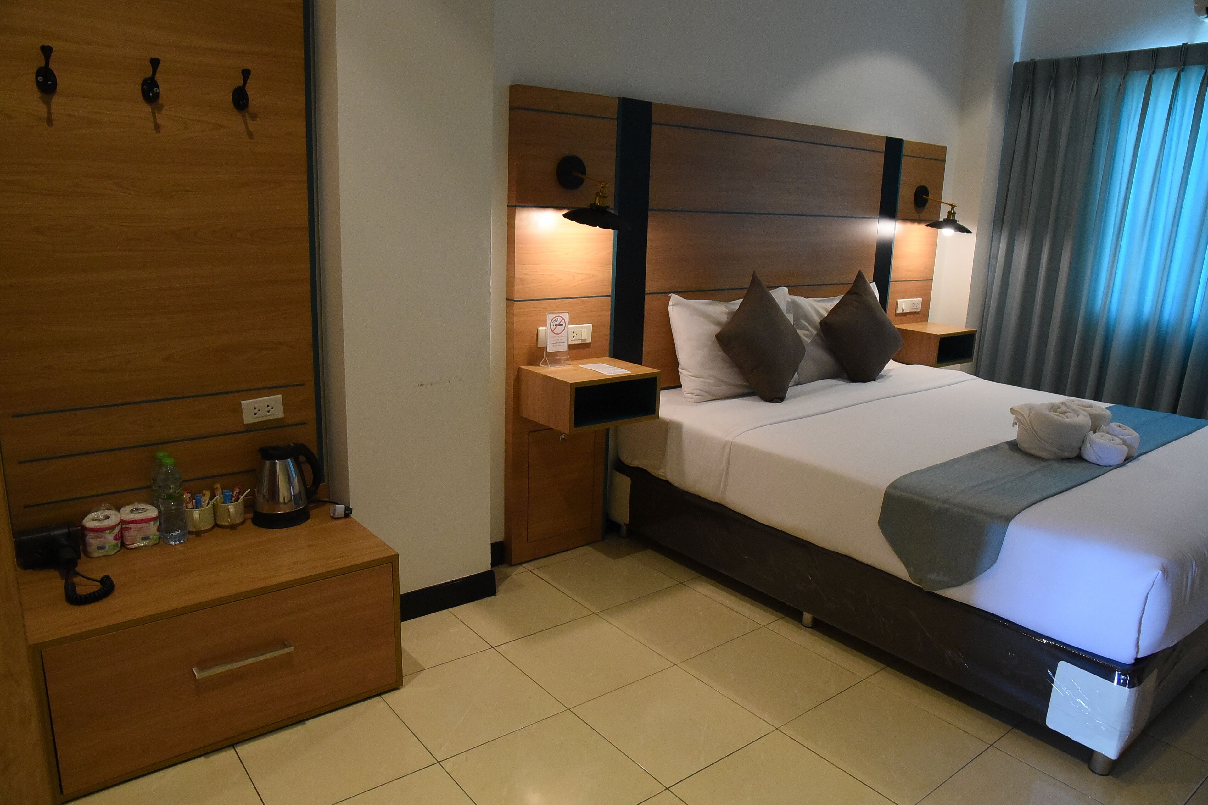 A hotel room with a wooden headboard, a bedside table with a lamp, a bed with pillows and a blanket, and a nightstand with a phone and a sign.