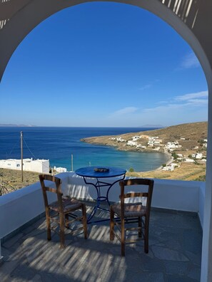 A view through an archway of a coastal area with a table and chairs, a clear sky, and distant buildings.