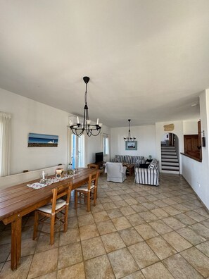A spacious dining area with a long wooden table and chairs, a chandelier, and a view of the sea through a window.