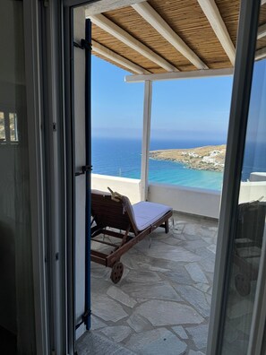 A balcony with a wooden lounge chair, a stone floor, and a view of the sea and buildings.