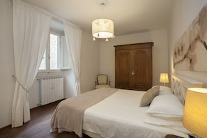 Un dormitorio con una cama grande, un armario de madera, una silla, un radiador y una ventana con cortinas.