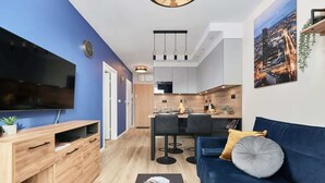 Una sala de estar moderna con un televisor de pantalla plana, mueble de entretenimiento de madera, sofá azul y un comedor con cocina al fondo.