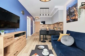 Una sala de estar moderna con un televisor de pantalla plana, mueble de entretenimiento de madera, sofá azul y un comedor con cocina al fondo.