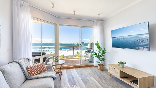 A modern living room with a large sliding glass door leading to a balcony, a sofa with cushions, a small table, a TV mounted on the wall, and a potted plant. - 203 The Breakwater, 2 Messines St - magical unit with lift, pool, views and aircon (Shoal Bay)