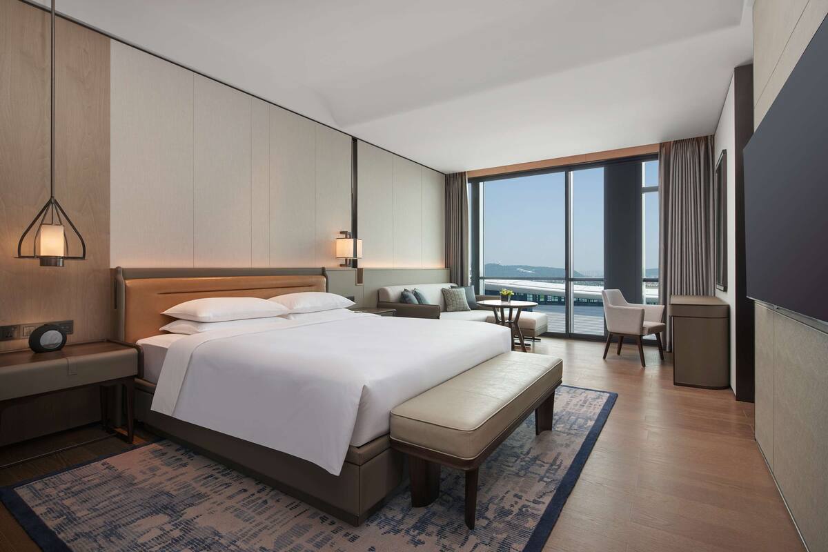 Habitación de hotel moderna con una cama grande, una mesita de noche, un banco, una pequeña mesa y vistas de la ciudad a través de amplias ventanas.