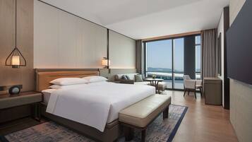 Habitación de hotel moderna con una cama grande, una mesita de noche, un banco, una pequeña mesa y vistas de la ciudad a través de amplias ventanas.