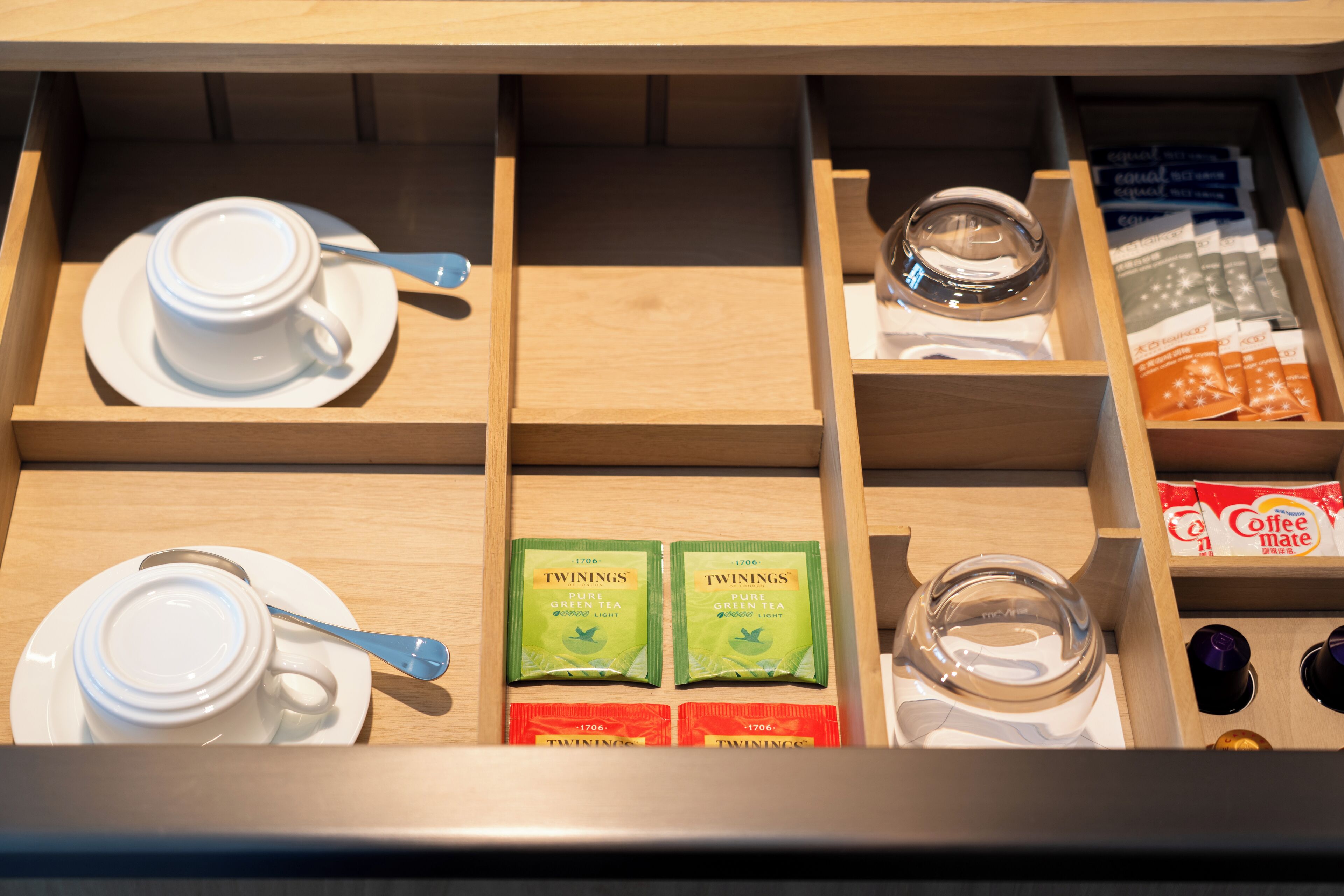 Un tiroir compartimenté contenant une tasse blanche, une cuillère, deux sachets de thé vert Twinings, un verre et un paquet de Coffee Mate.