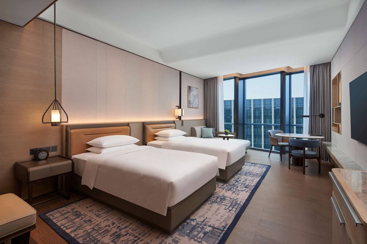 Habitación de hotel con dos camas, un escritorio, una silla, un televisor y un ventanal con vistas.