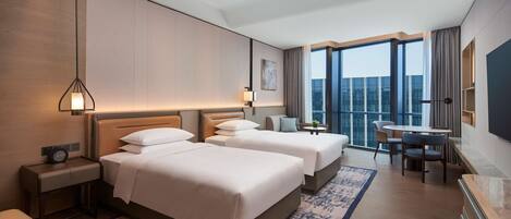 A hotel room with two beds, a desk, a chair, a TV, and a large window with a view.