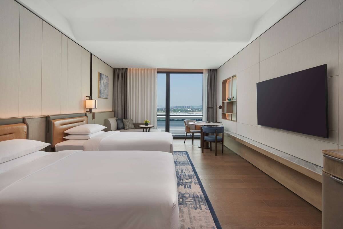 Habitación de hotel con dos camas, un escritorio, una silla, un televisor y vistas a la ciudad.