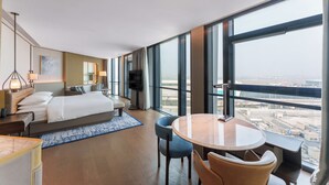 Habitación de hotel moderna con una cama grande, un escritorio, una silla, una mesa redonda y un televisor. La habitación tiene ventanas amplias que ofrecen vistas de la ciudad.