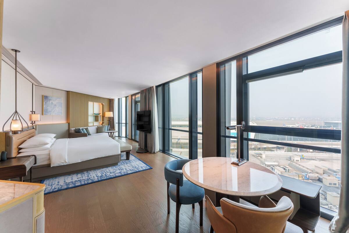Habitación de hotel moderna con una cama grande, un escritorio, una silla, una mesa redonda y un televisor. La habitación tiene ventanas amplias que ofrecen vistas de la ciudad.