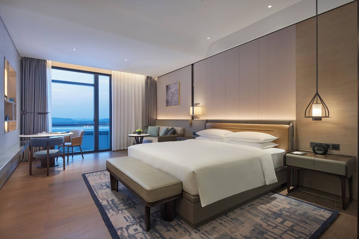 Una habitación de hotel moderna con una cama grande, un escritorio, una silla, un banco y vistas al exterior.