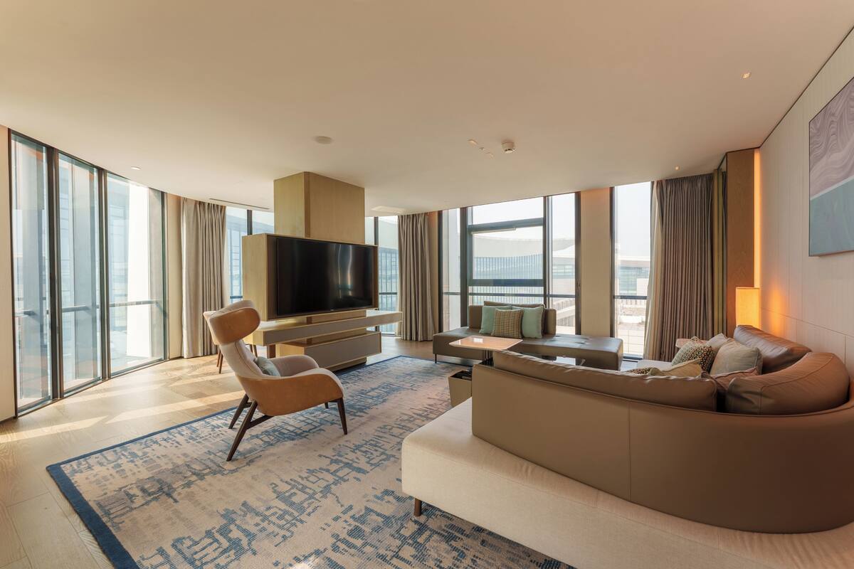 Una habitación de hotel moderna con un ventanal grande, un televisor de pantalla plana, un sofá, una silla y una mesa de centro.