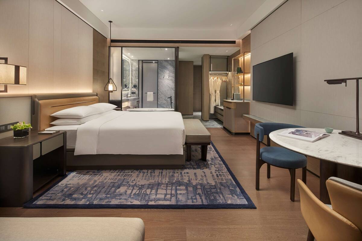 Una habitación de hotel moderna con una cama grande, mesitas de noche, un escritorio con una silla, un televisor y un baño visible a través de una puerta corredera.